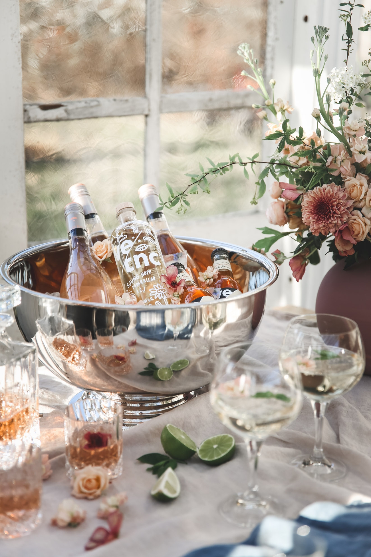 Vinkylare & Champagnekylare fylld med flaskor på bord med vit duk samt rosa blommor och drinkar