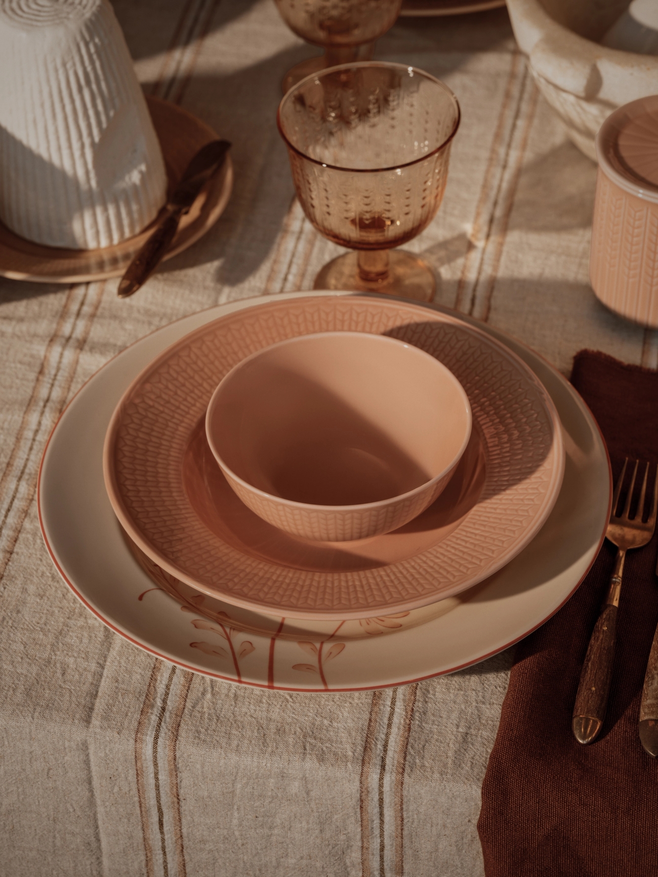 A rustic table setting with stacked pink and beige dishes, amber glassware, a striped linen tablecloth, and ceramic containers.