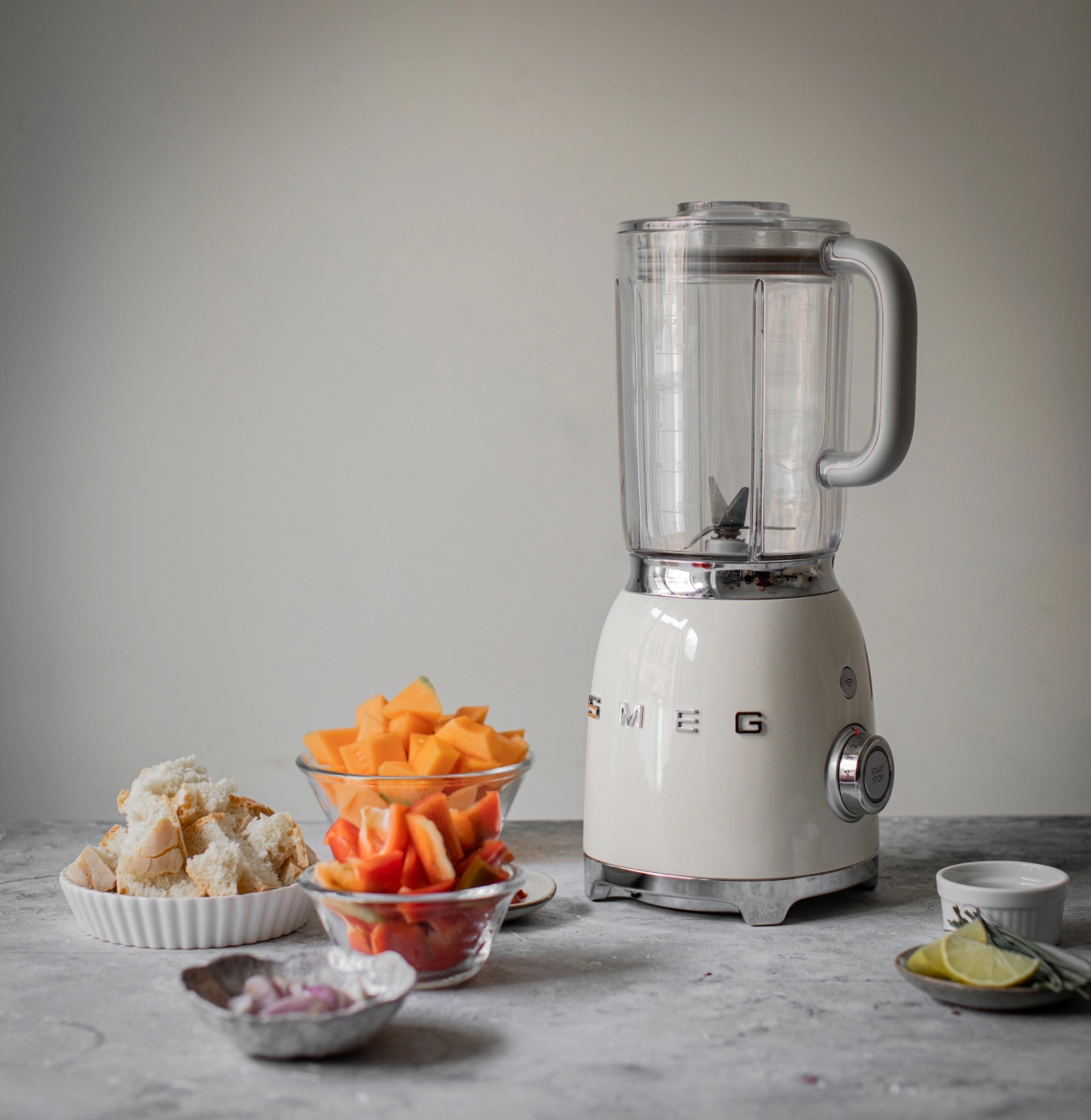 A cream-colored Smeg blender on a counter surrounded by bowls of diced pumpkin, red peppers, bread, and chopped onions, with lime and herbs.