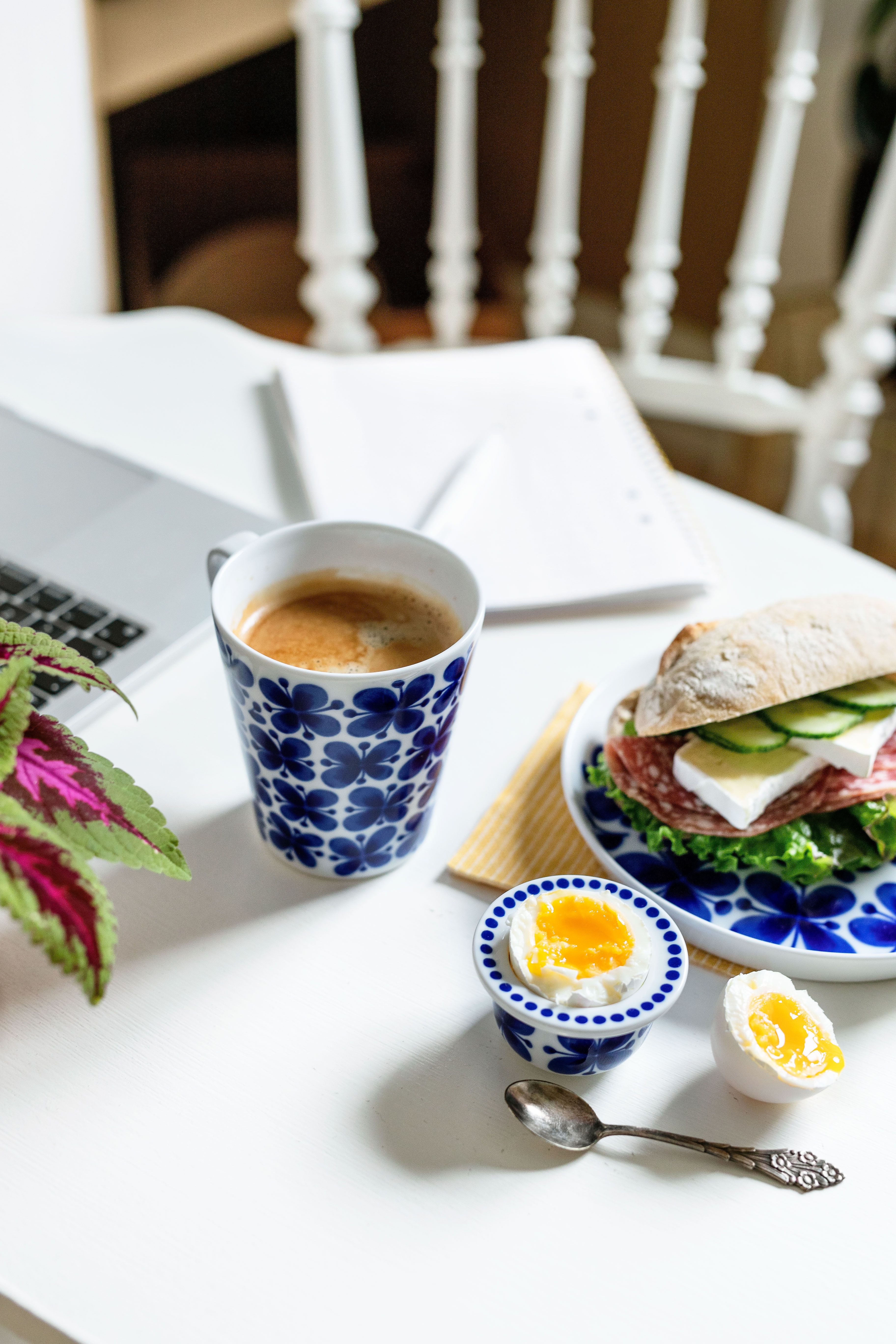 Ett vitt bord dukat med kaffe i en blåmönstrad mugg, en smörgås, löskokta ägg, en bärbar dator och en växt.