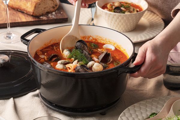 En hånd holder en svart støpejernsgryte fylt med sjømatlapskaus, inkludert blåskjell, muslinger og reker.