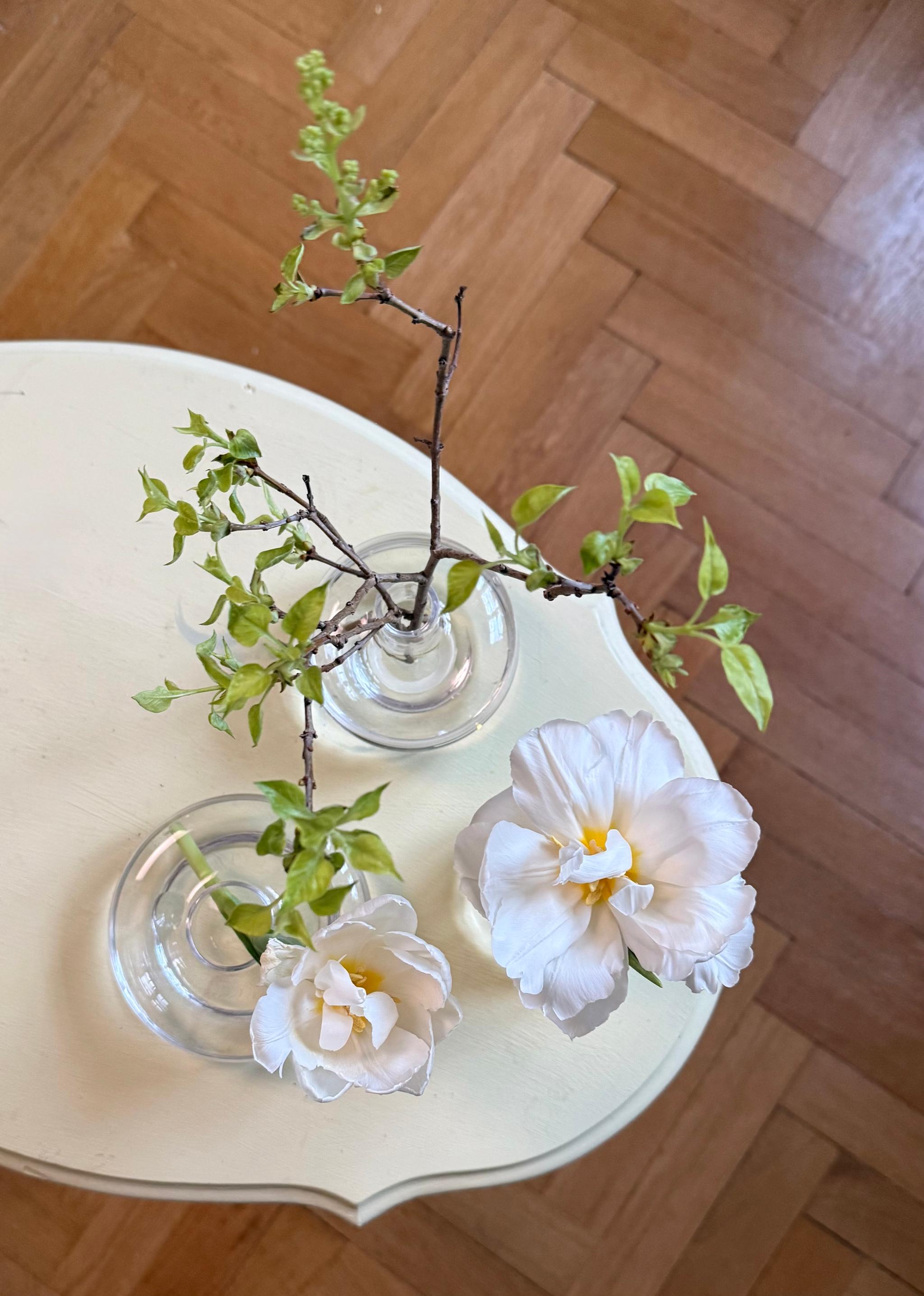 Two white tulips and two budding green branches in small glass vases on a light-colored table, viewed from above.