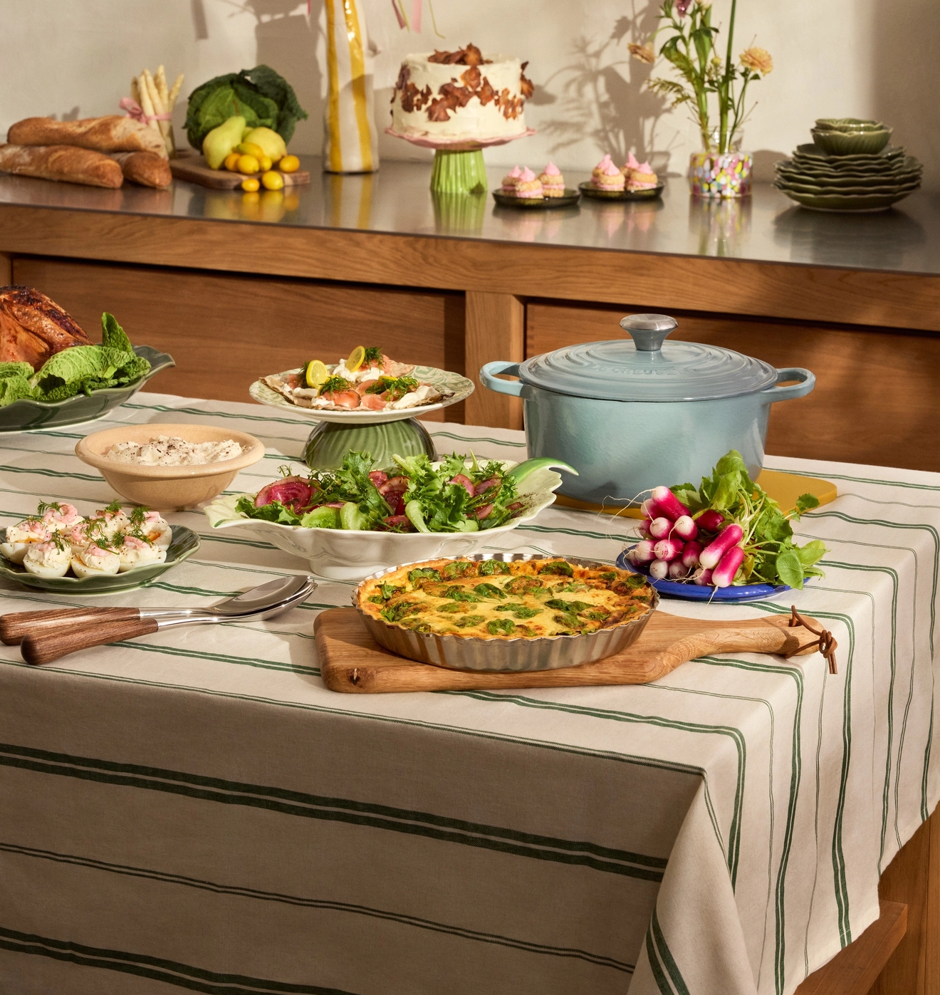 Ett matbord dukat med en grönrandig duk är fyllt med en paj, sallader, fyllda ägghalvor och en ljusblå gryta, med en tårta, bröd och blomsterdekorationer på en bänk i bakgrunden