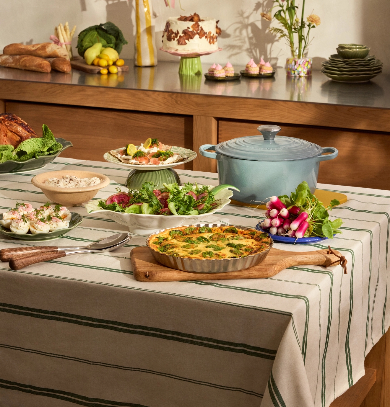Et festlig måltid servert på en stripete duk og på en kjøkkenbenk, med pai, salater, brød, kake og vårdekorasjoner.