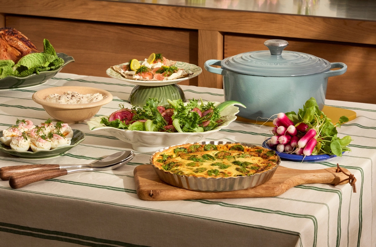 Ett matbord dukat med en grönrandig duk är fyllt med en paj, sallader, fyllda ägghalvor och en ljusblå gryta, med en tårta, bröd och blomsterdekorationer på en bänk i bakgrunden