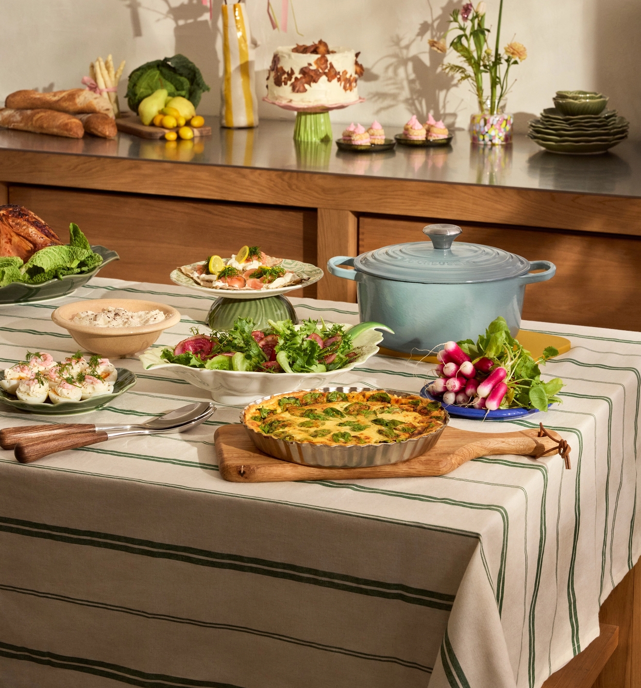 Juhlava ruokapöytä ja keittiötaso ovat täynnä keväistä ateriaa, kuten piirakkaa, salaatteja, leipää, kakkua ja kukkakoristeita