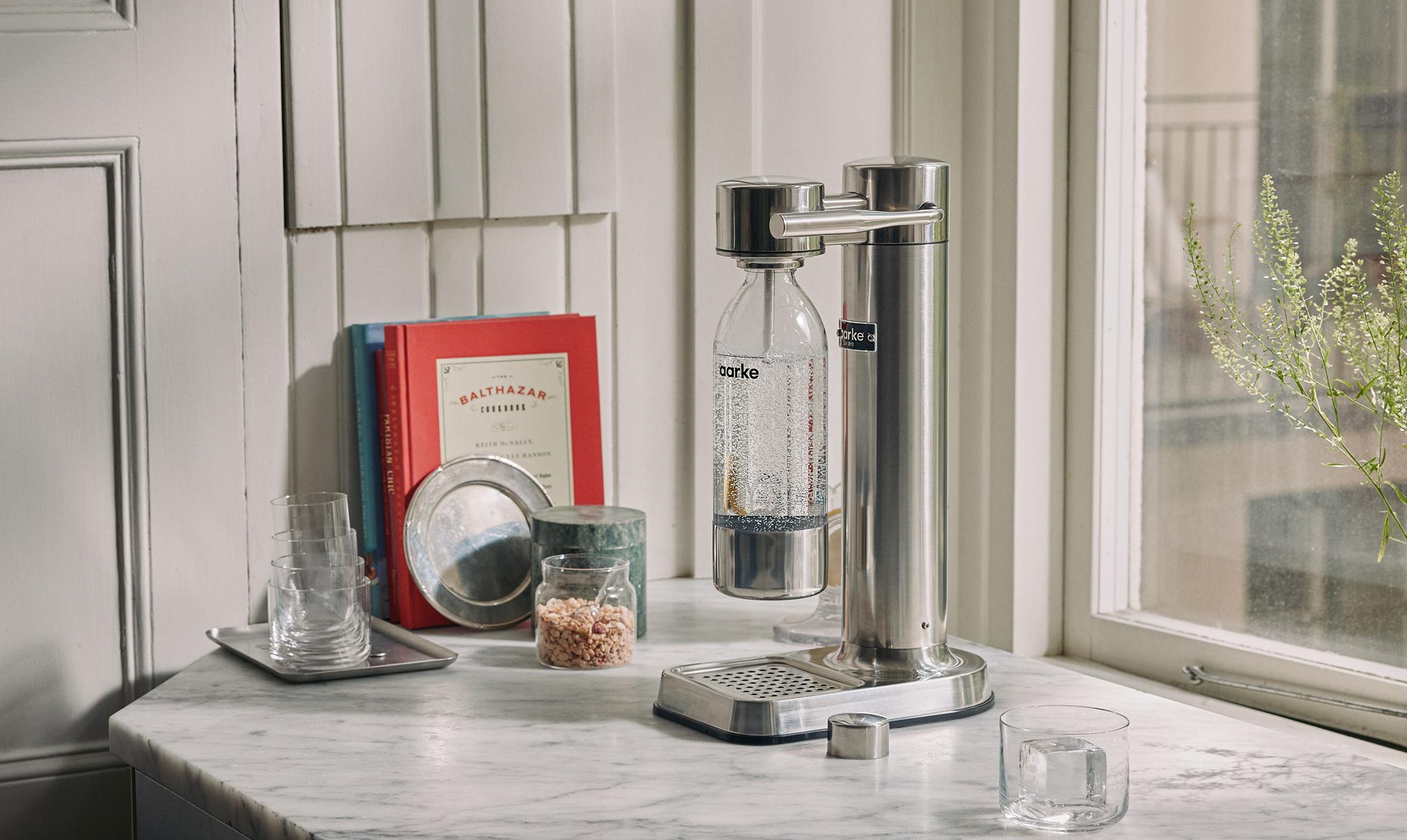 Modern köksbänk i marmor med kolsyremaskin, glas och kokbok vid ett solbelyst fönster.