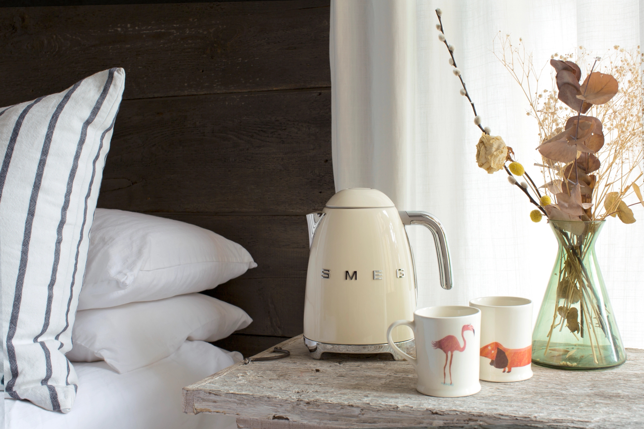 Cream Smeg kettle, flamingo mug, dog mug, and a vase of dried flowers on a rustic bedside table.