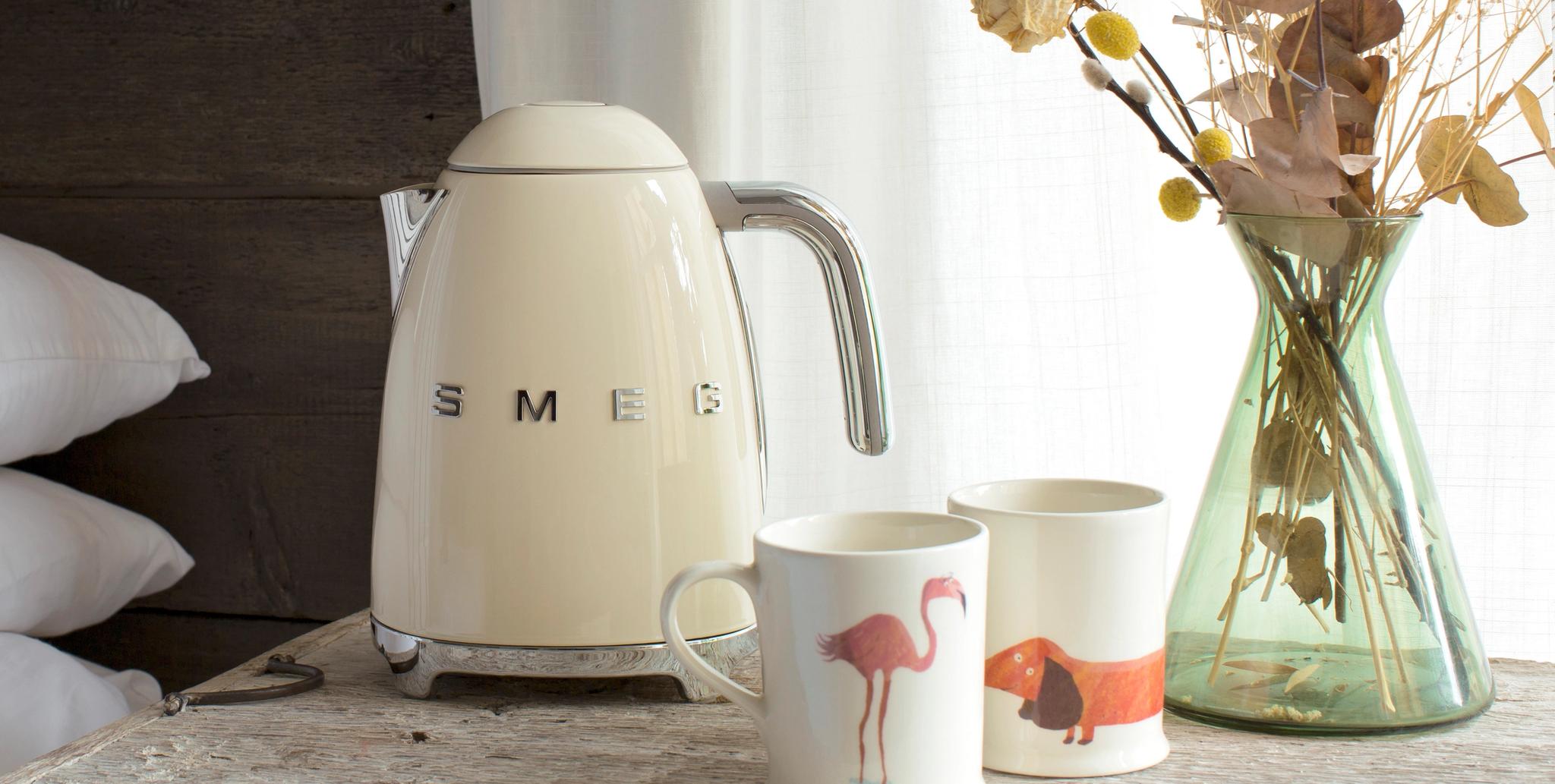 Kermanvärinen Smeg-vedenkeitin kromikahvalla, pinottujen valkoisten tyynyjen ja valkoisen mukin vieressä.