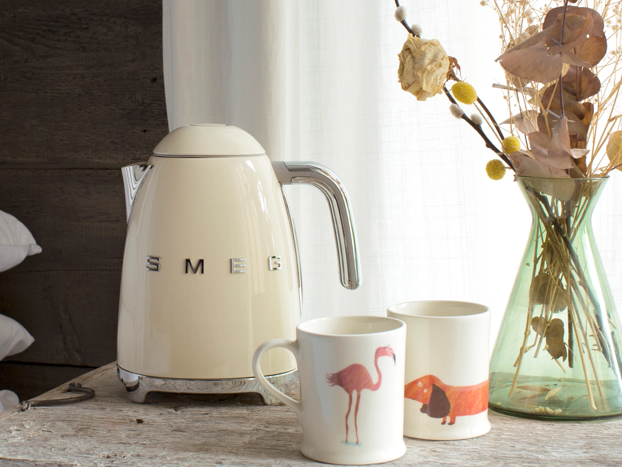 Kermanvärinen Smeg-vedenkeitin kromikahvalla, pinottujen valkoisten tyynyjen ja valkoisen mukin vieressä.