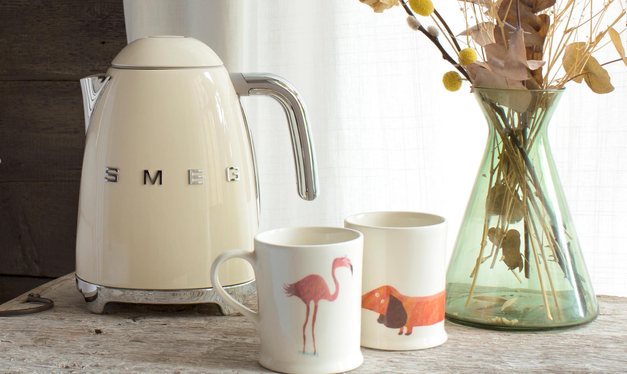 En krämfärgad Smeg-vattenkokare på en rustik trähylla bredvid vita kuddar.