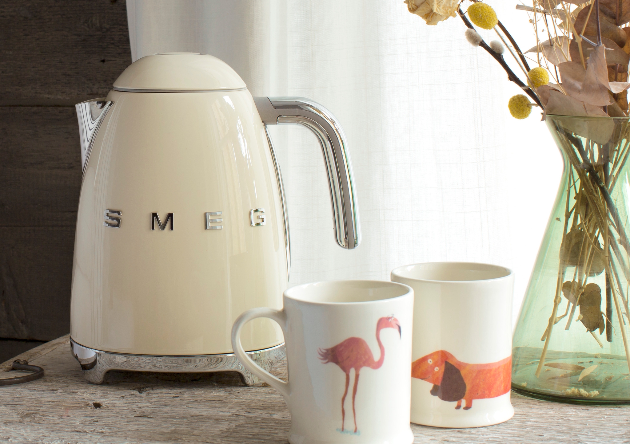 En krämfärgad Smeg-vattenkokare på en rustik trähylla bredvid vita kuddar.