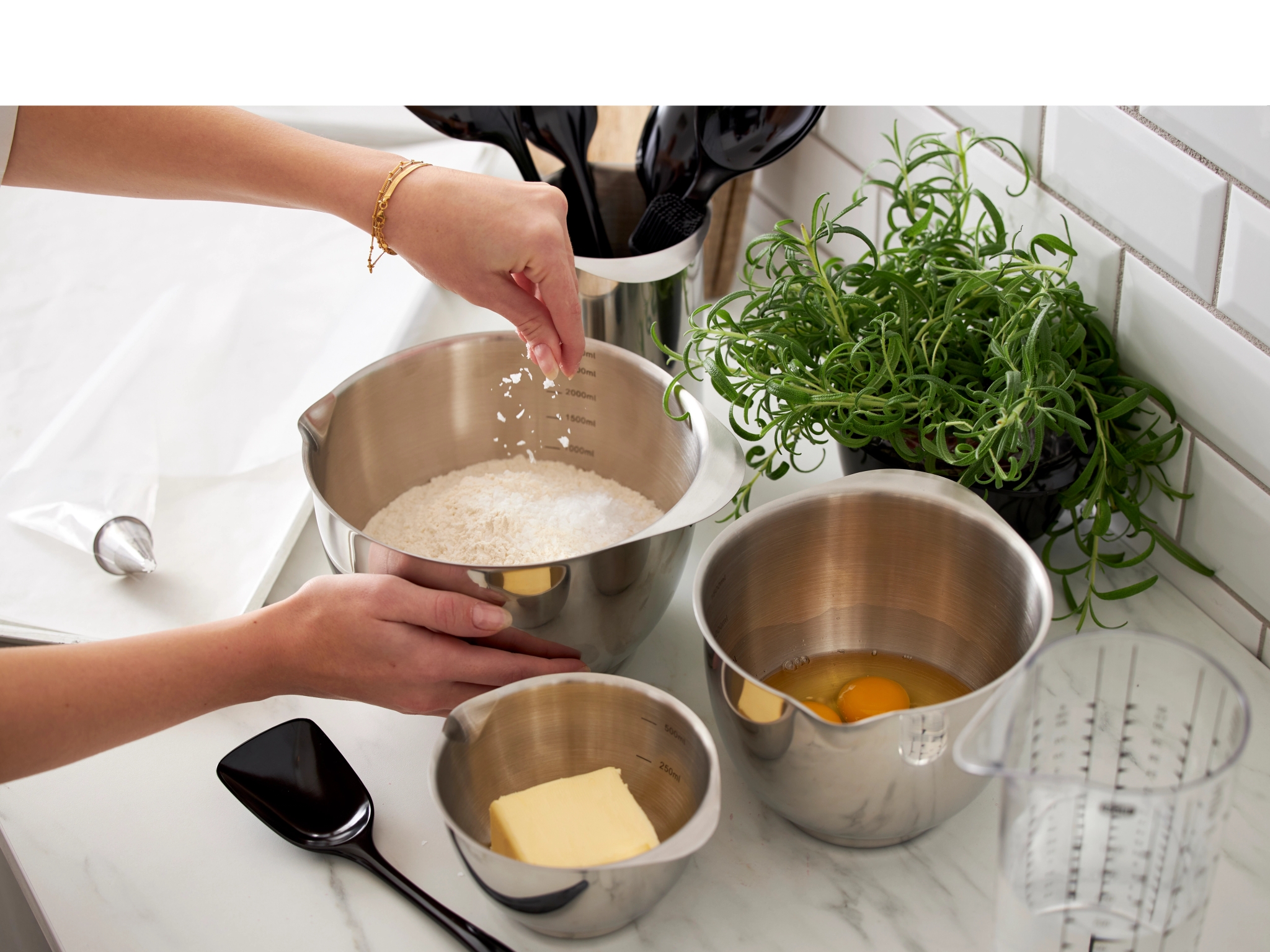 Henkilö ripottelee ainesosan suureen ruostumattomasta teräksestä valmistettuun kulhoon, ja keittiön tiskillä on pienempiä kulhoja, joissa on voita ja munia.