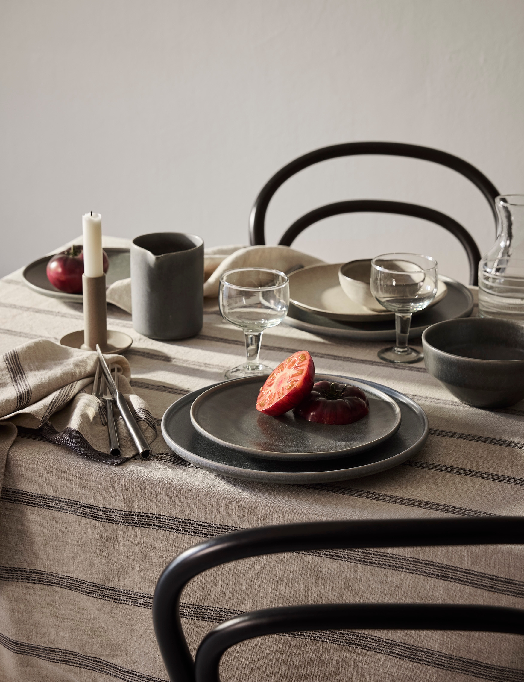 Ett matbord dukat med rustika keramiktallrikar, glas, bestick och ett ljus på en randig linneduk, med skivade tomater på en tallrik och mörka stolar