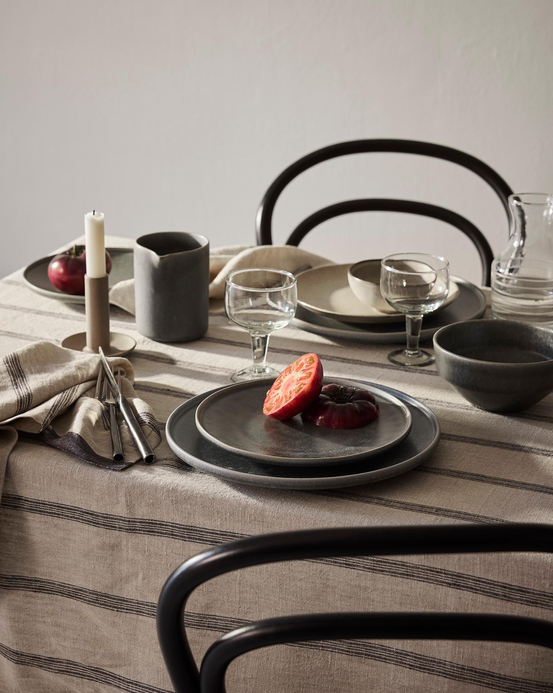 Ett matbord dukat med rustika keramiktallrikar, glas, bestick och ett ljus på en randig linneduk, med skivade tomater på en tallrik och mörka stolar