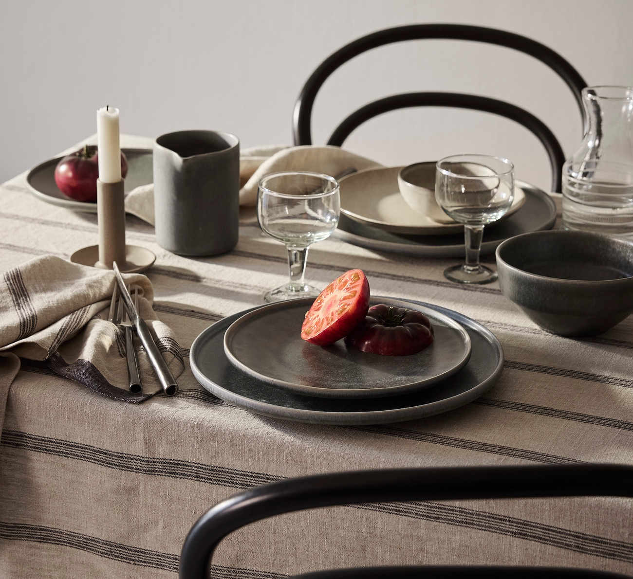 Ett matbord dukat med rustika keramiktallrikar, glas, bestick och ett ljus på en randig linneduk, med skivade tomater på en tallrik och mörka stolar