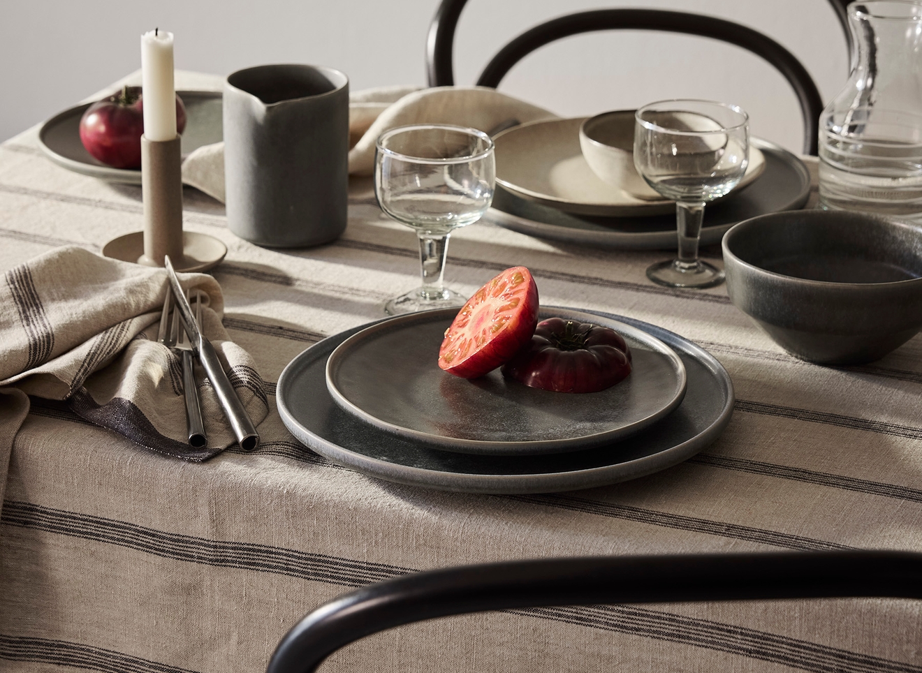 Ett matbord dukat med rustika keramiktallrikar, glas, bestick och ett ljus på en randig linneduk, med skivade tomater på en tallrik och mörka stolar