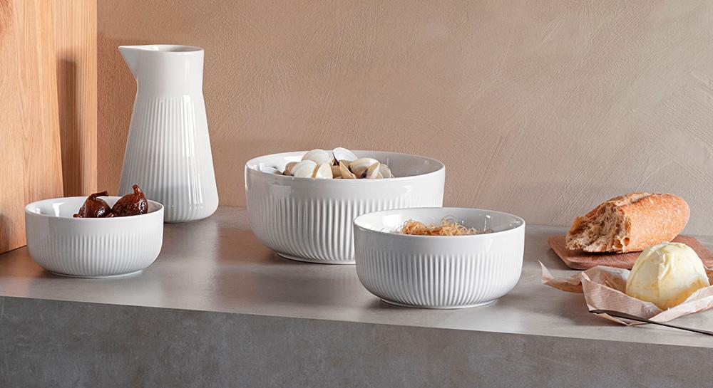 Valkoisia uurrettuja kulhoja, joissa on viikunoita, simpukoita ja kuivattuja nuudeleita, sekä valkoinen maljakko.