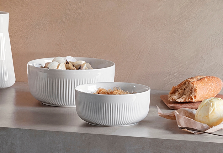 Valkoisia uurrettuja kulhoja, joissa on viikunoita, simpukoita ja kuivattuja nuudeleita, sekä valkoinen maljakko.