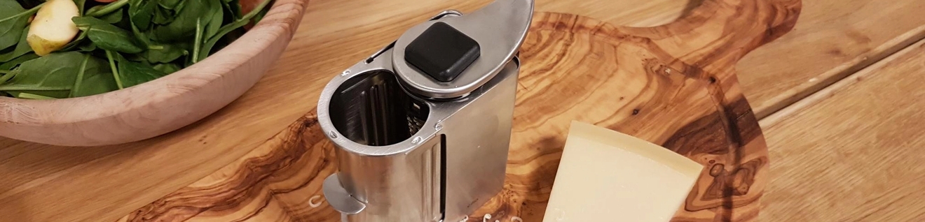 Metallinen juustoraastin, raastettua juustoa ja juustokimpaleita puutarjoilulaudalla, taustalla vihreä salaatti.