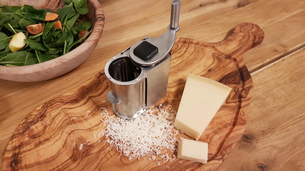 Metallinen juustoraastin, raastettua juustoa ja juustokimpaleita puutarjoilulaudalla, taustalla vihreä salaatti.