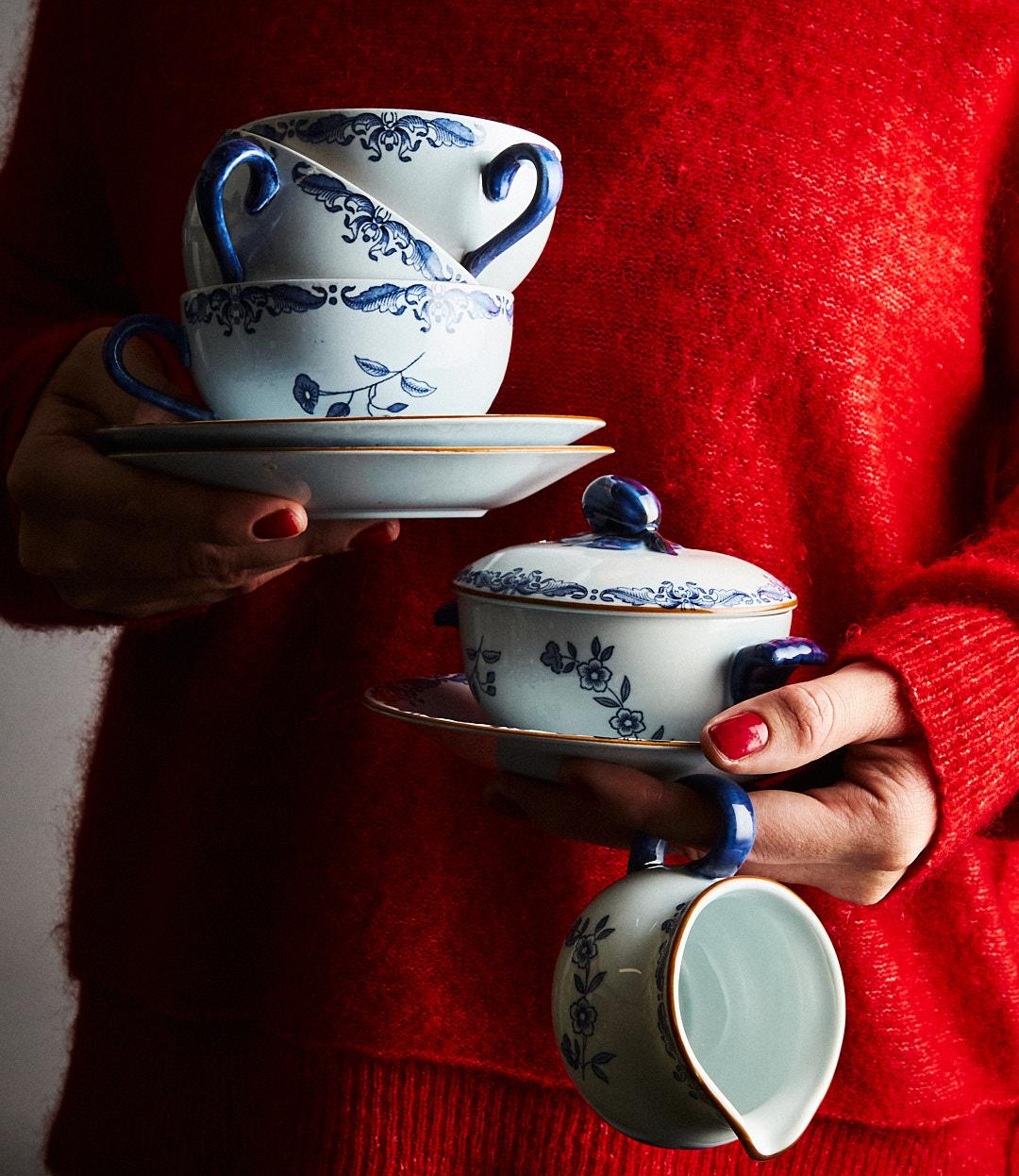 En person i en röd tröja håller blå- och vitblommönstrade koppar, fat, en skål med lock och en gräddkanna