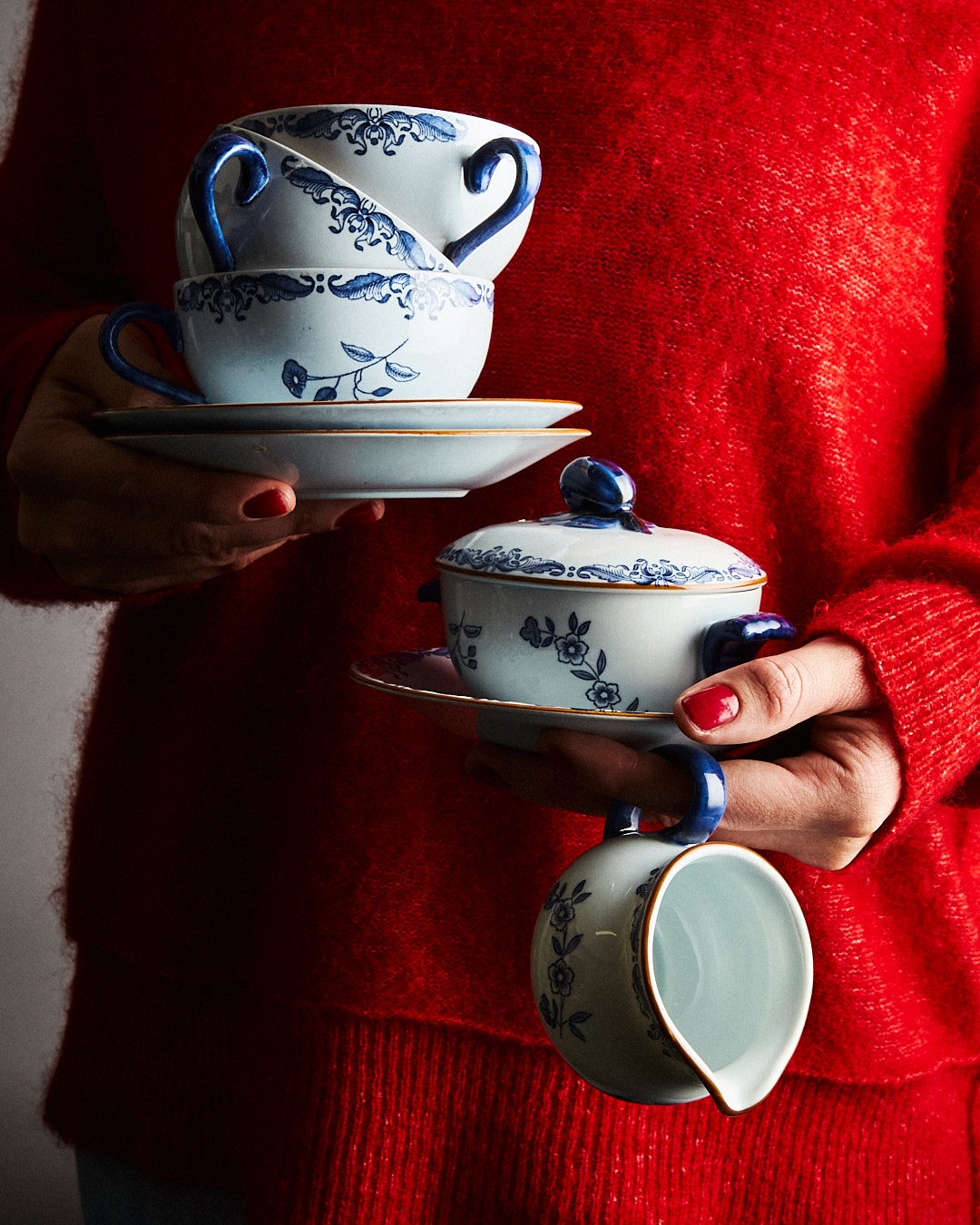 En person i en röd tröja håller blå- och vitblommönstrade koppar, fat, en skål med lock och en gräddkanna