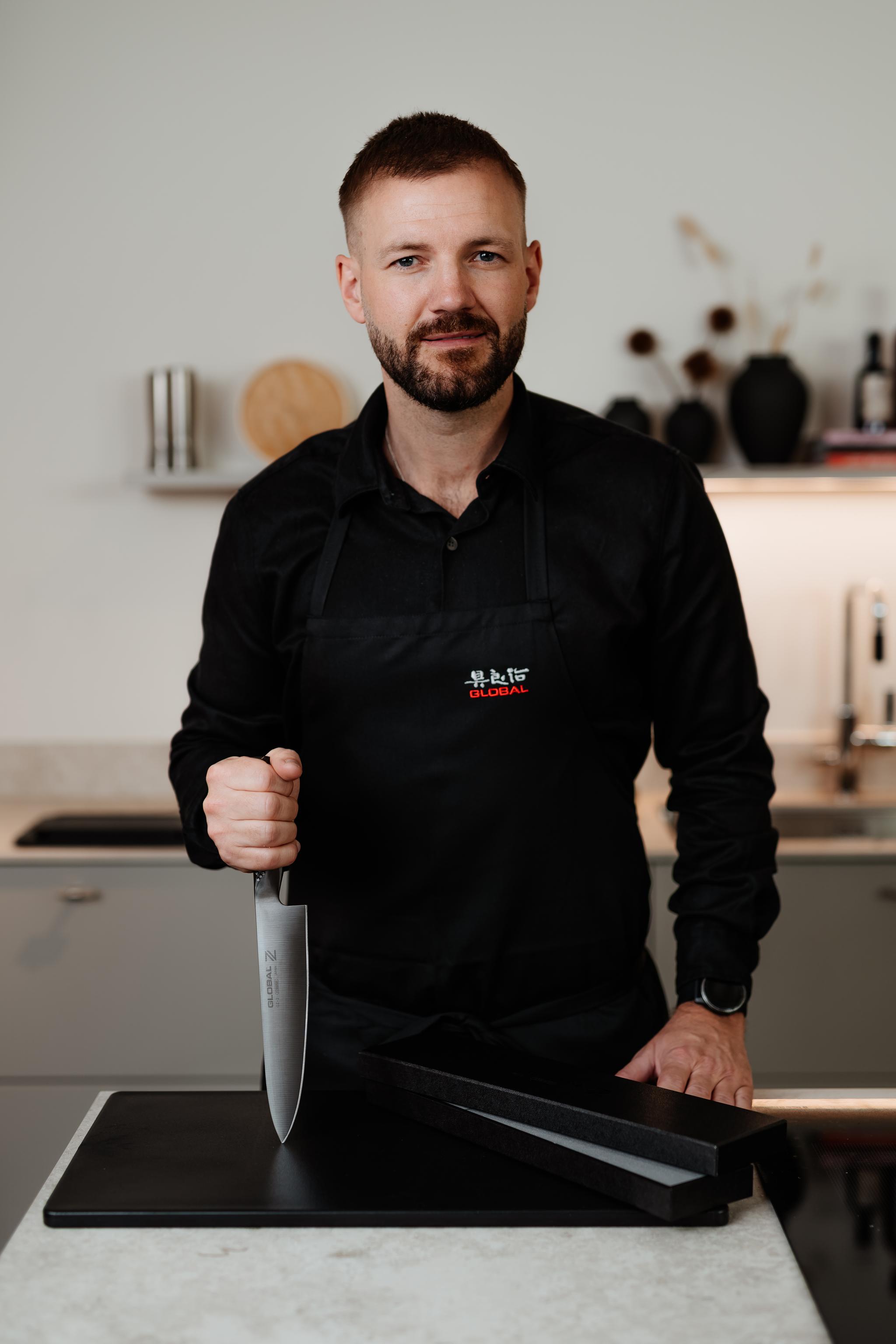 En man i ett svart förkläde håller en stor kockkniv med spetsen vilande på en svart skärbräda i ett modernt kök.