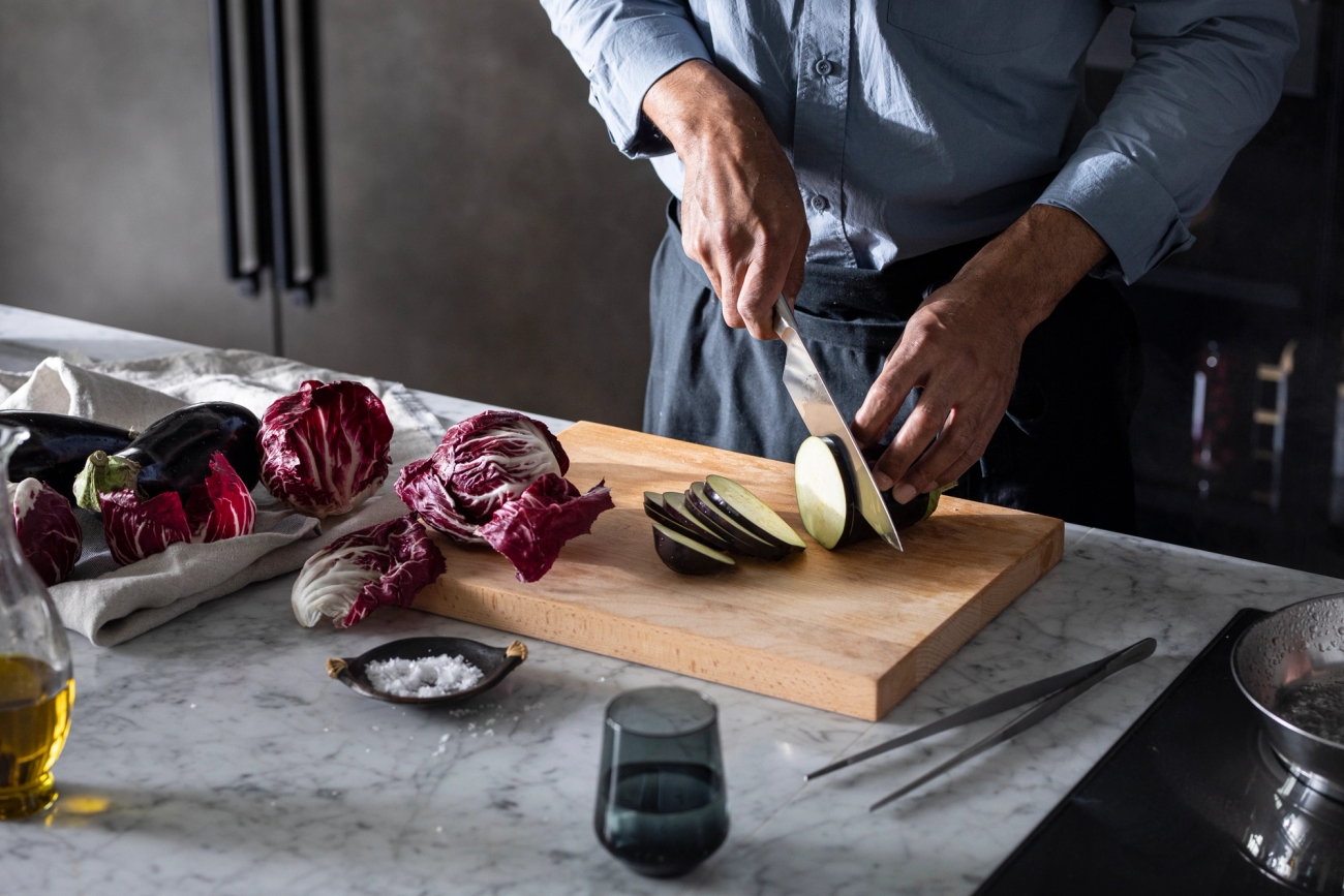 Person skär aubergine på en skärbräda i ett modernt kök med radicchio och olivolja på marmorbänken.