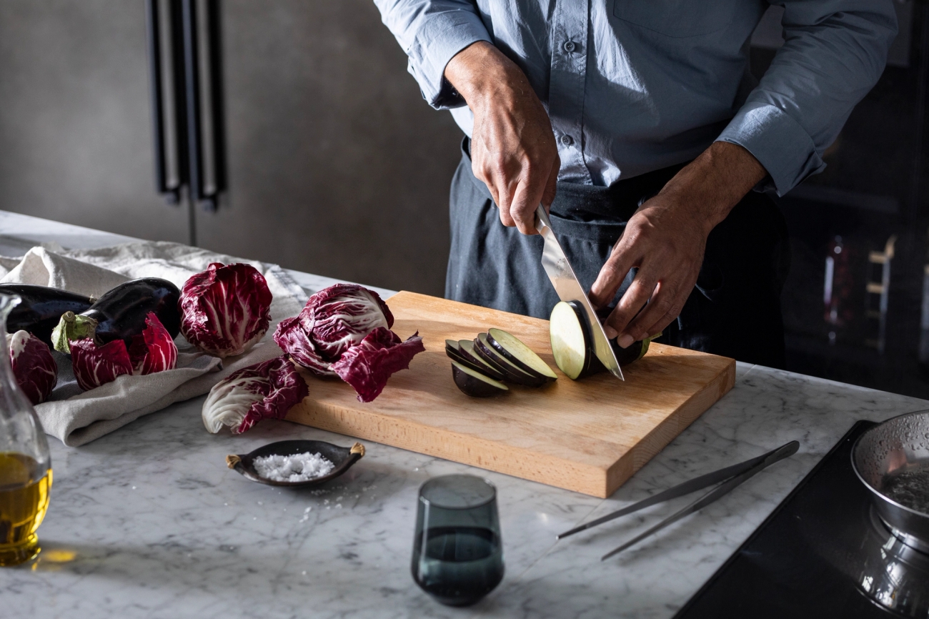 Person skär aubergine på en skärbräda i ett modernt kök med radicchio och olivolja på marmorbänken.