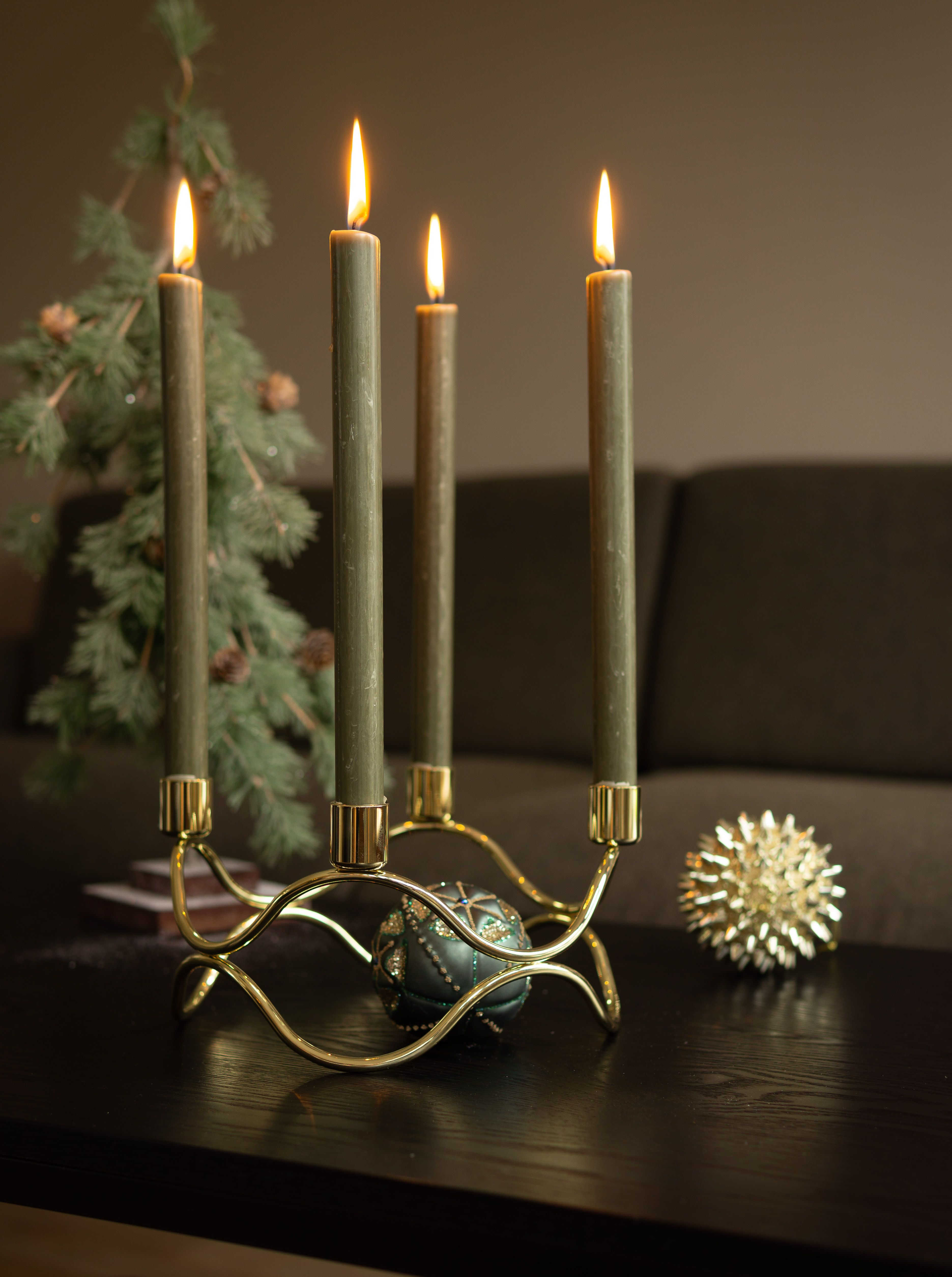 Kultainen kynttelikkö, jossa palaa neljä vihreää kynttilää, taustalla sumeita joulukoristeita.
