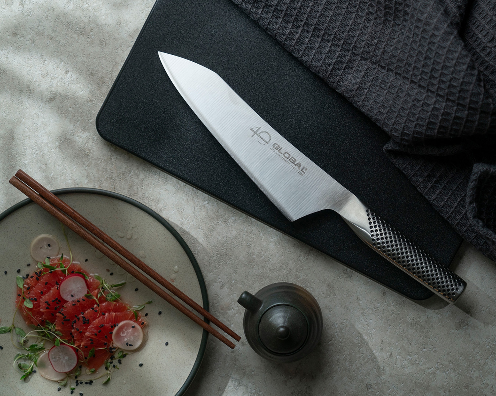 Global-kockkniv på svart skärbräda bredvid sashimi, ätpinnar och en liten tekanna.