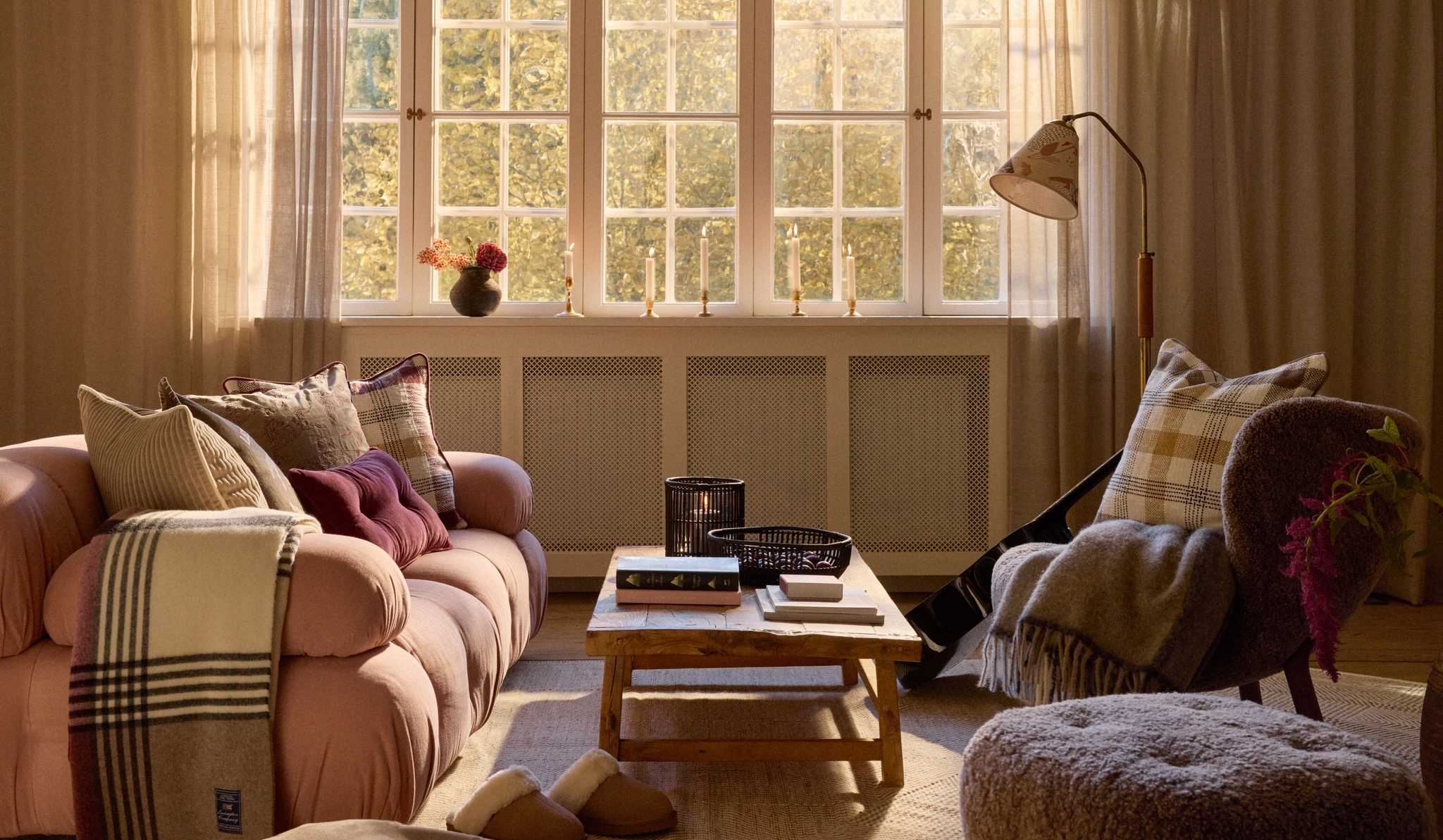 Solbelyst vardagsrum med rosa soffa, mjuka textilier, fåtölj och soffbord framför stora spröjsade fönster.