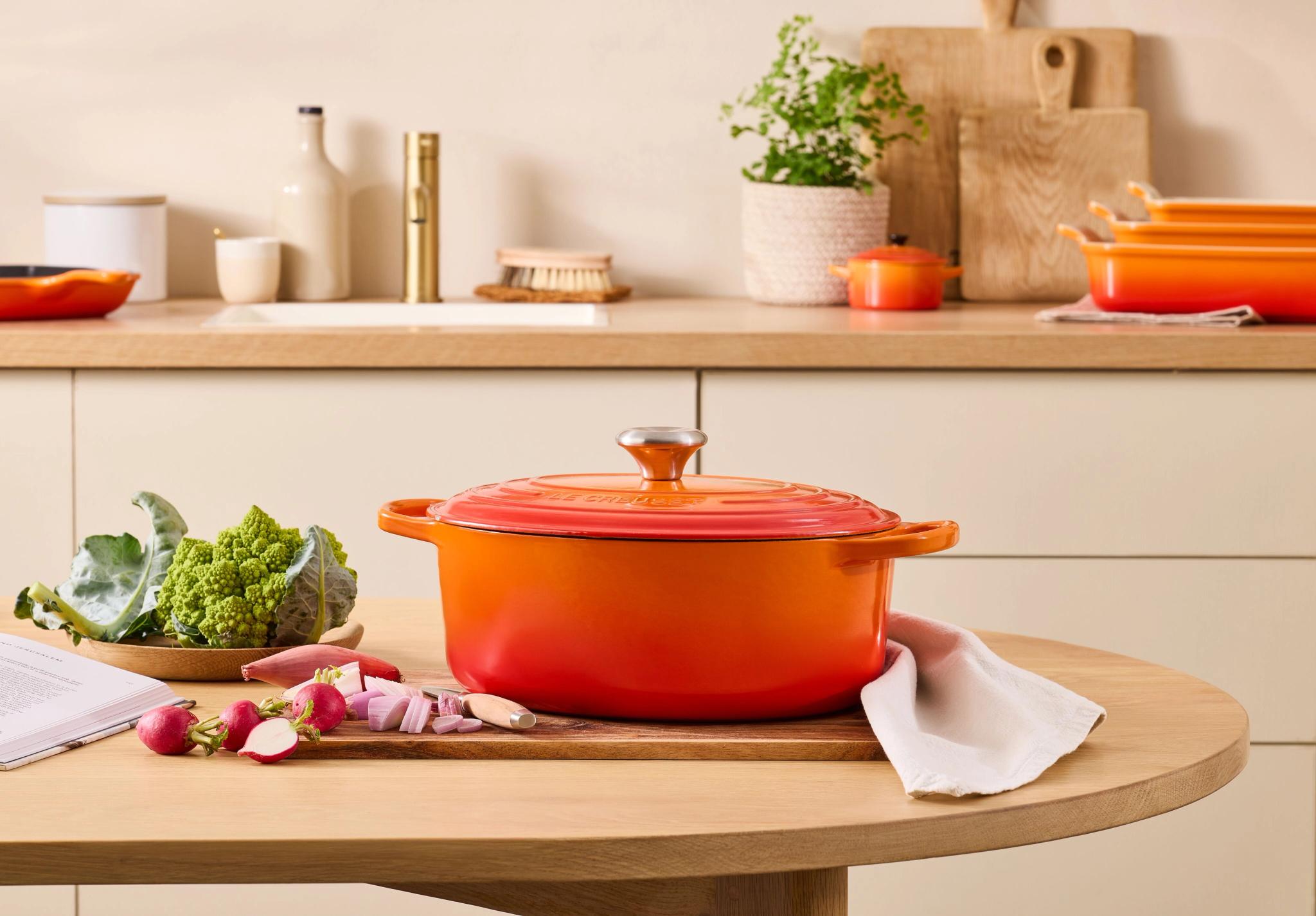 Orange Le Creuset-gryta på ett träbord med grönsaker, en kokbok och en köksbakgrund.