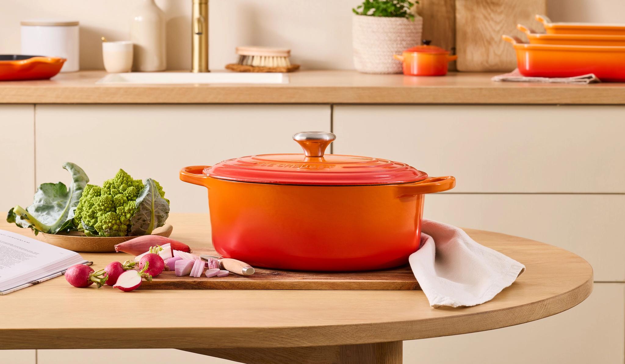 Orange Le Creuset-gryta på ett träbord med grönsaker, en kokbok och en köksbakgrund.