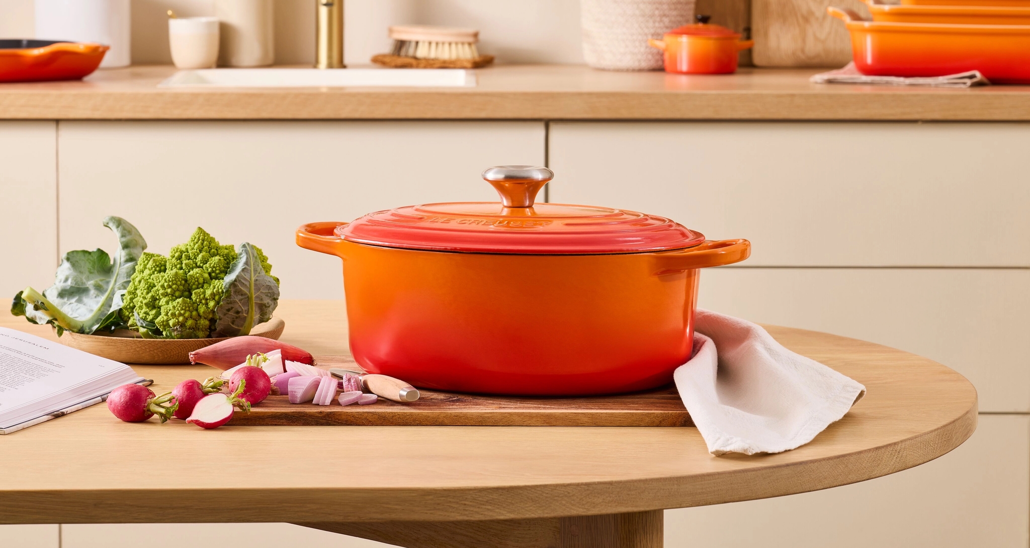 Kirkkaan oranssi Le Creuset -kattila puisella keittiönpöydällä, jonka päällä on tuoreita vihanneksia ja yhteensopivia keittiövälineitä sen takana olevalla tiskillä.
