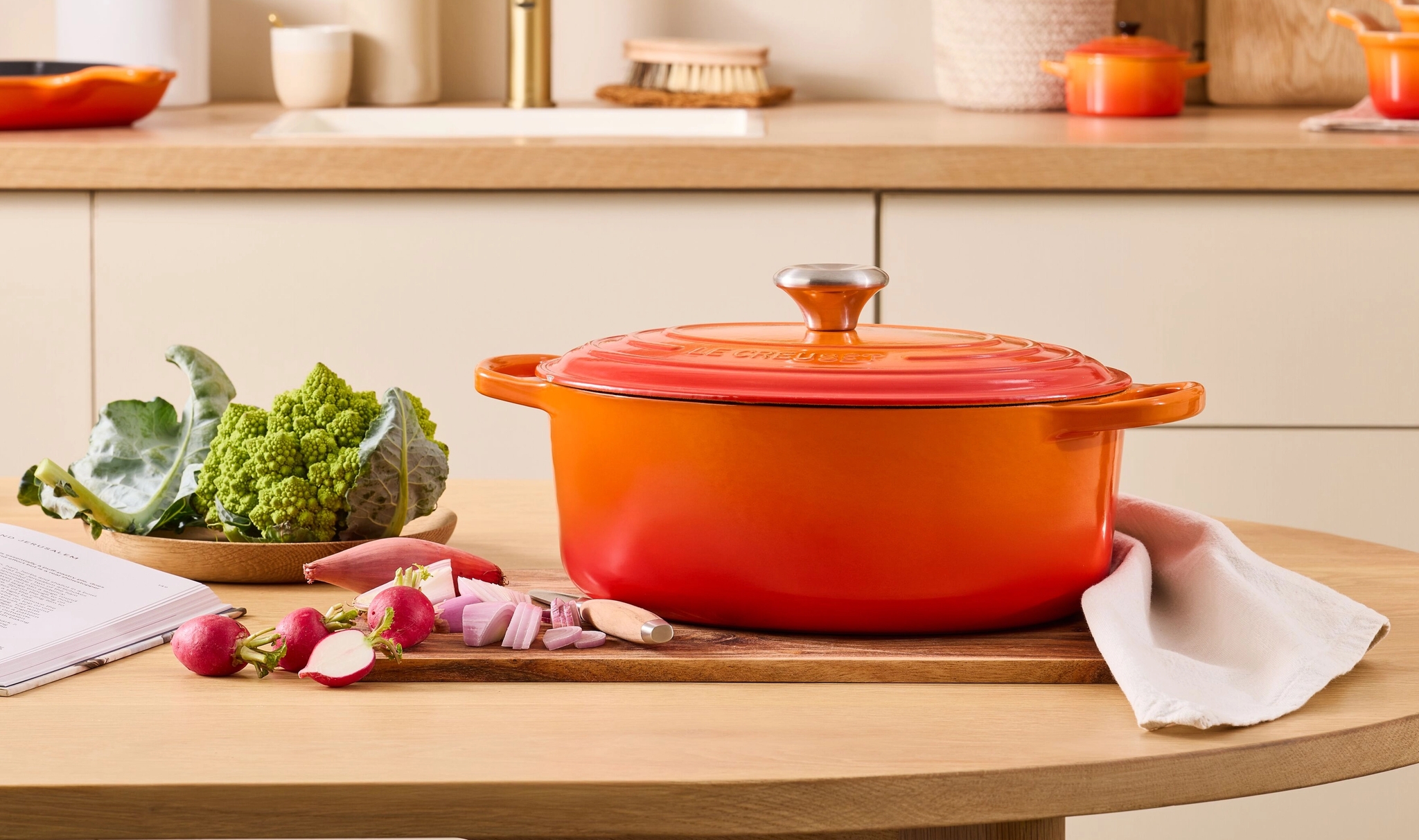 Orange Le Creuset-gryta på ett träbord med grönsaker, en kokbok och en köksbakgrund.
