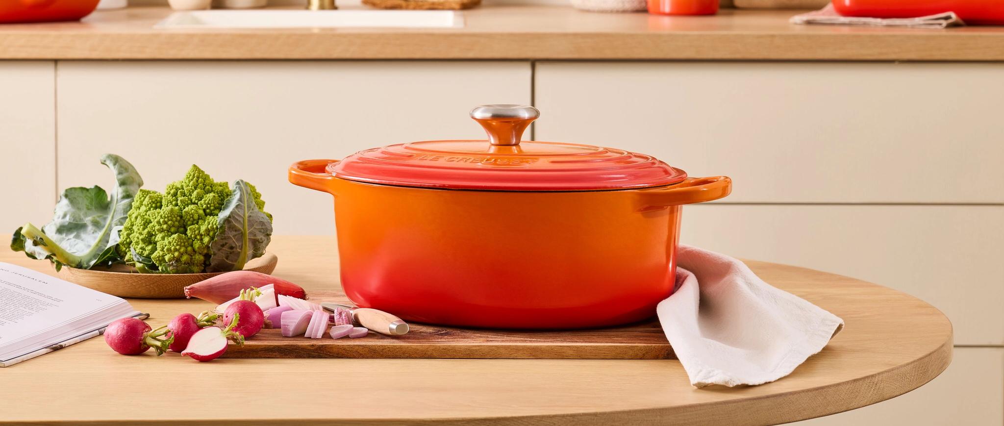 Kirkkaan oranssi Le Creuset -kattila puisella keittiönpöydällä, jonka päällä on tuoreita vihanneksia ja yhteensopivia keittiövälineitä sen takana olevalla tiskillä.