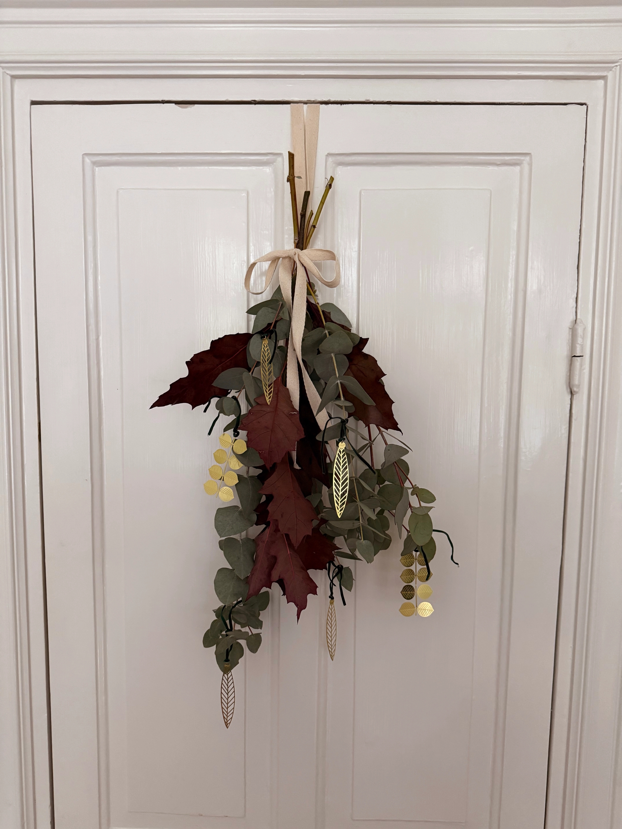 Autumnal swag of eucalyptus, red leaves, and gold metallic ornaments tied with a ribbon, hanging on a white door.