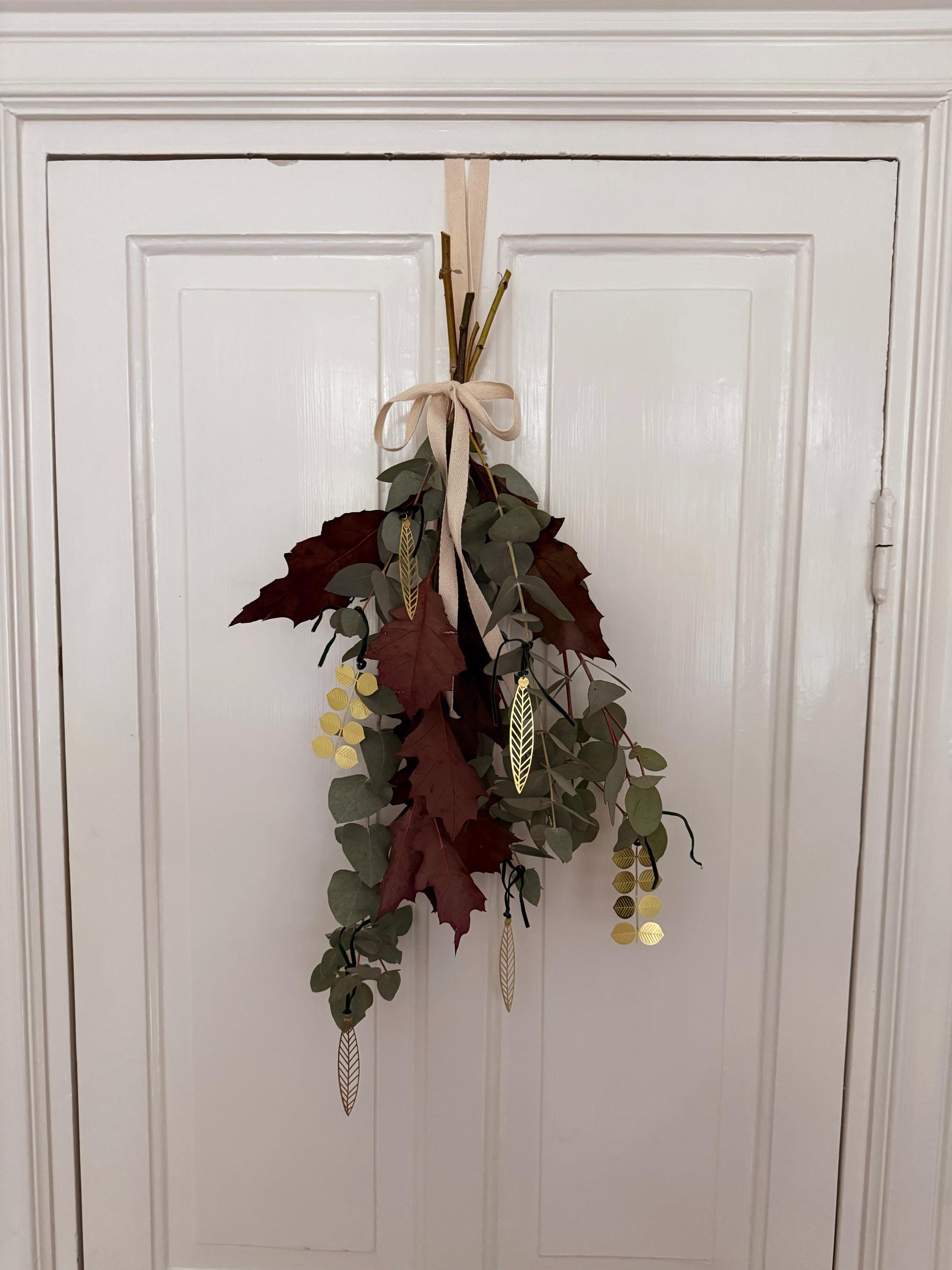 Decorative door swag with green eucalyptus, burgundy leaves, beige ribbon, and gold ornaments on a white door.
