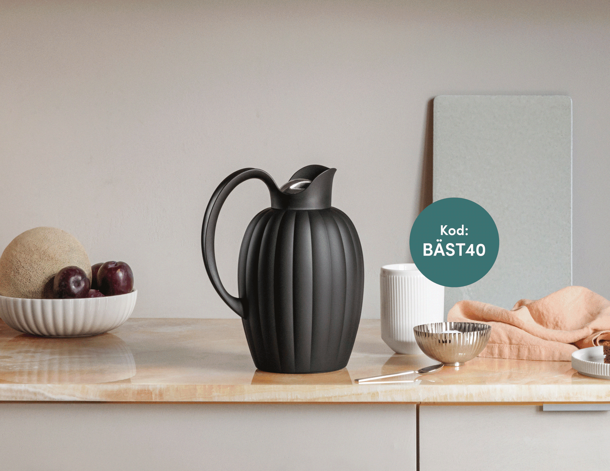 En svart räfflad termos på en bänk med en skål med frukt, en vit mugg och en silverskål.