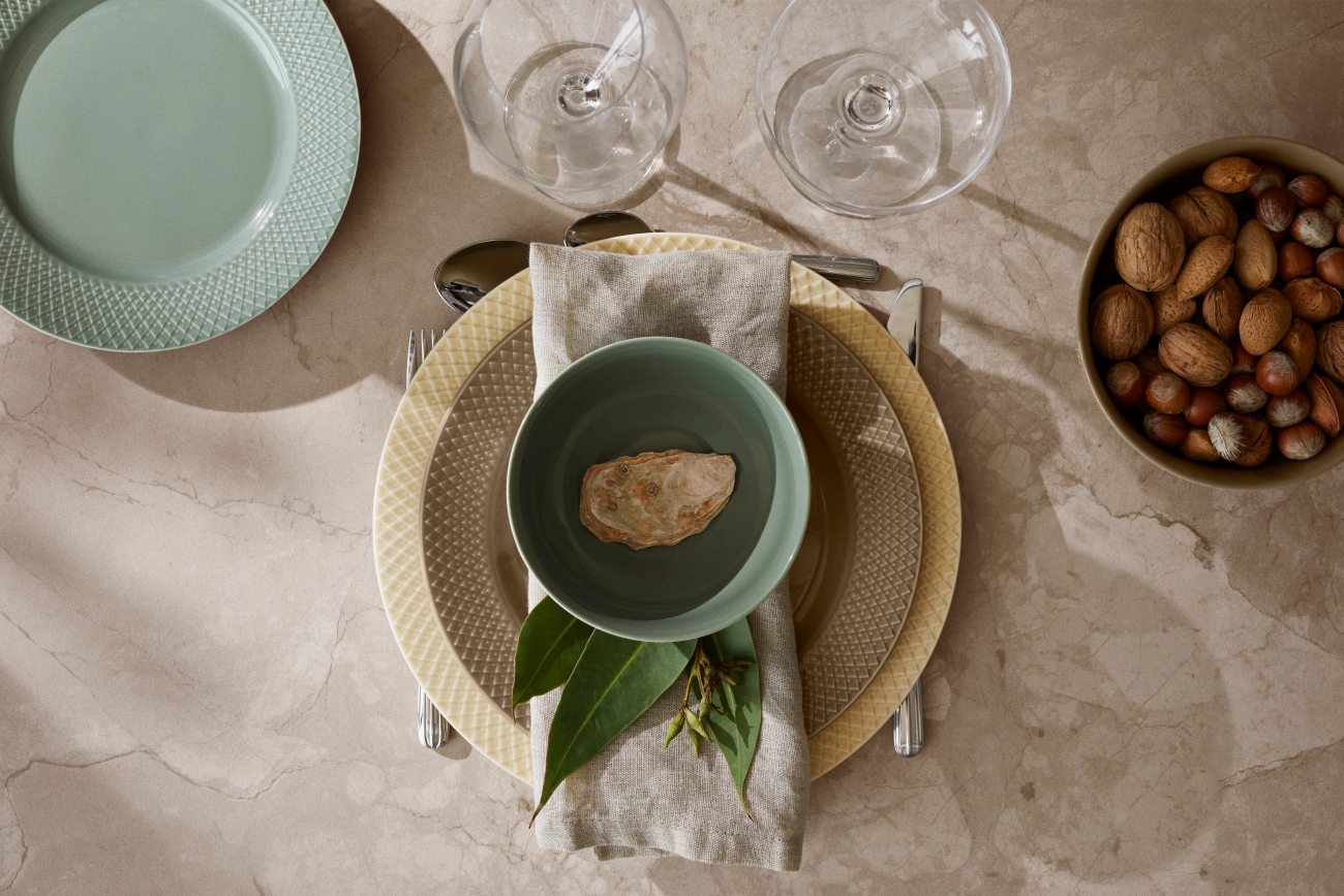 Ovanifrånvy av ett matbord dukat med texturerade tallrikar, en grön skål med en dekorativ sten, eukalyptusblad, bestick, vinglas och en skål med nötter på en solig marmoryta.