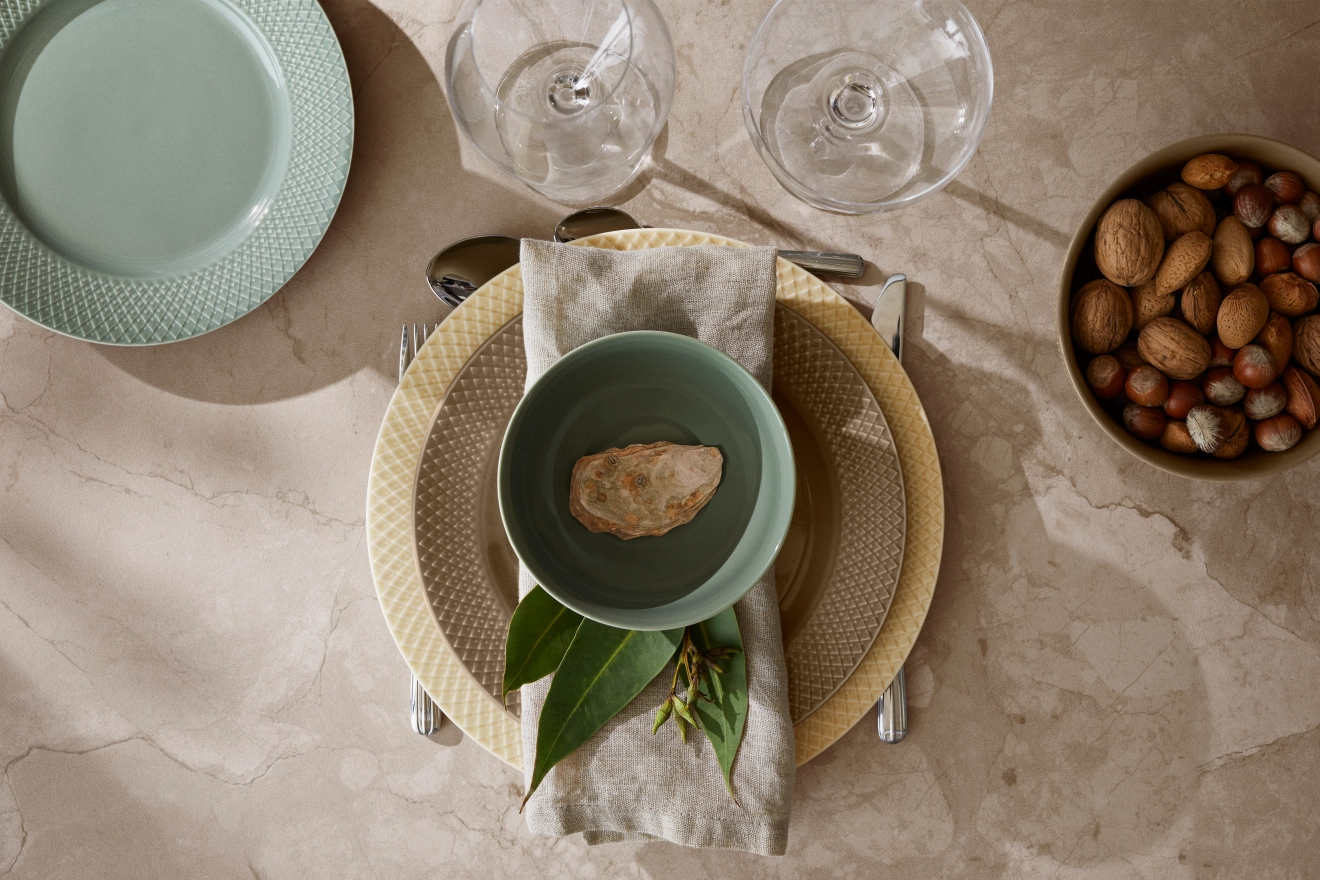 Ovanifrånvy av ett matbord dukat med texturerade tallrikar, en grön skål med en dekorativ sten, eukalyptusblad, bestick, vinglas och en skål med nötter på en solig marmoryta.
