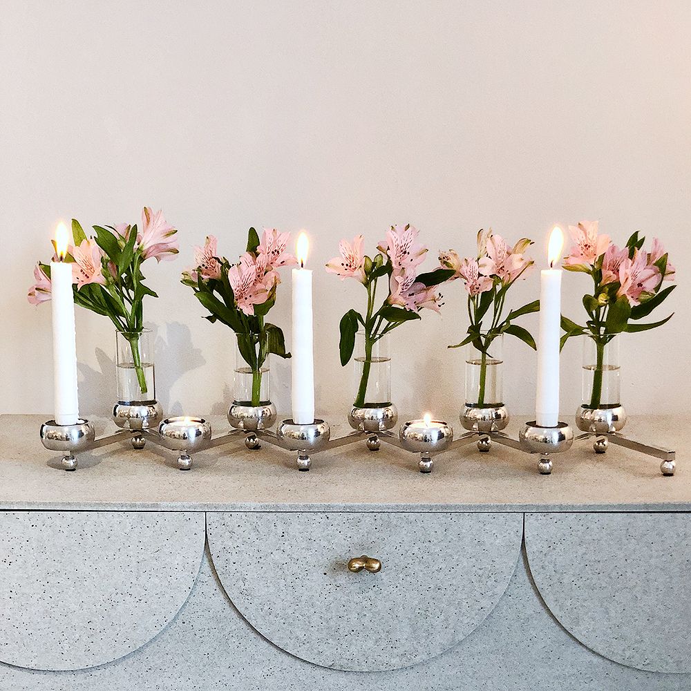 En polerad silverkandelaber med tända vita ljus och rosa alstroemerior i vaser står på en gråspräcklig byrå.