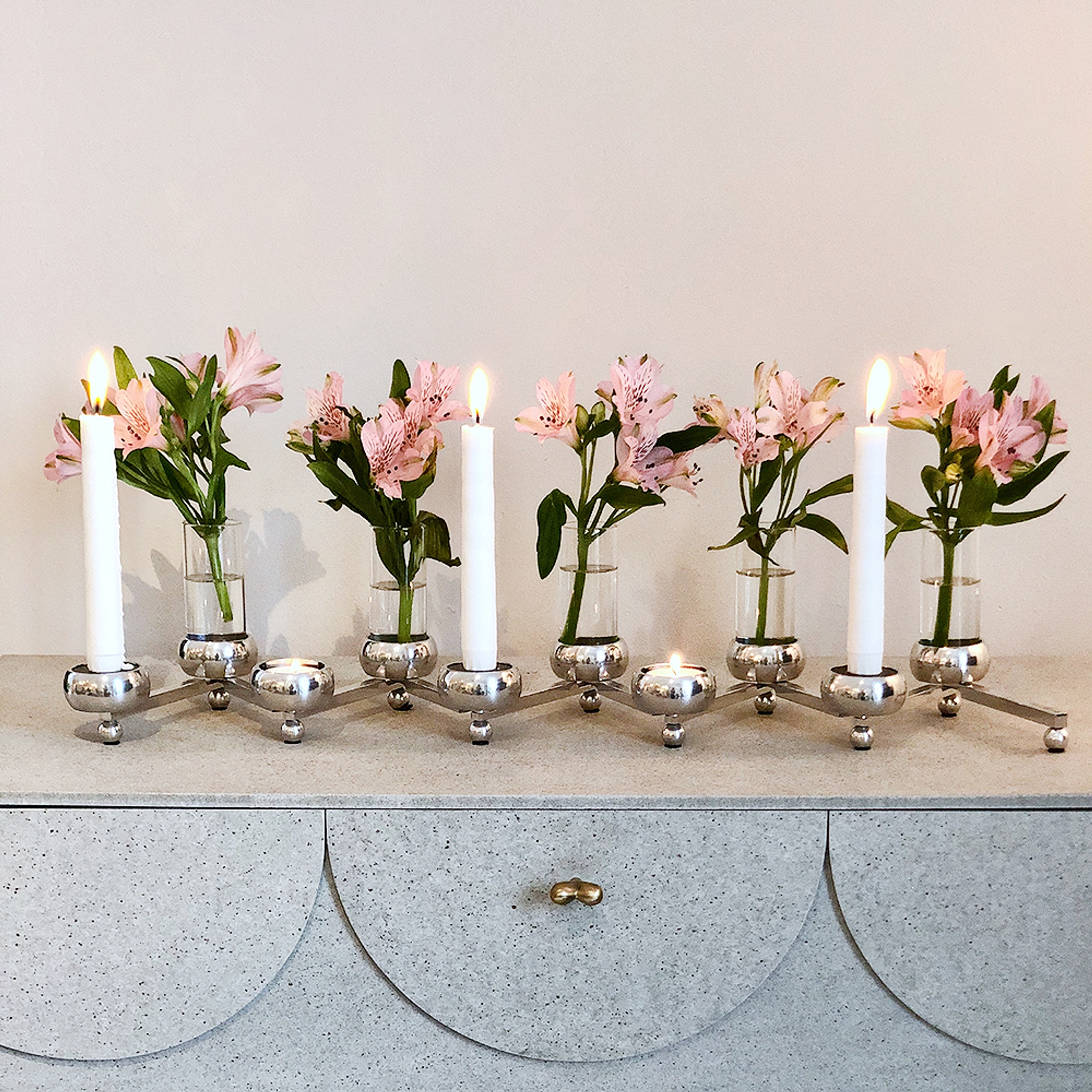 Moderni hopeinen kynttelikkö, jossa on kolme palavaa kynttilää ja pieniä vaaleanpunaisia ​​kukkia sisältävä maljakoita vaaleanharmaalla lipastolla.