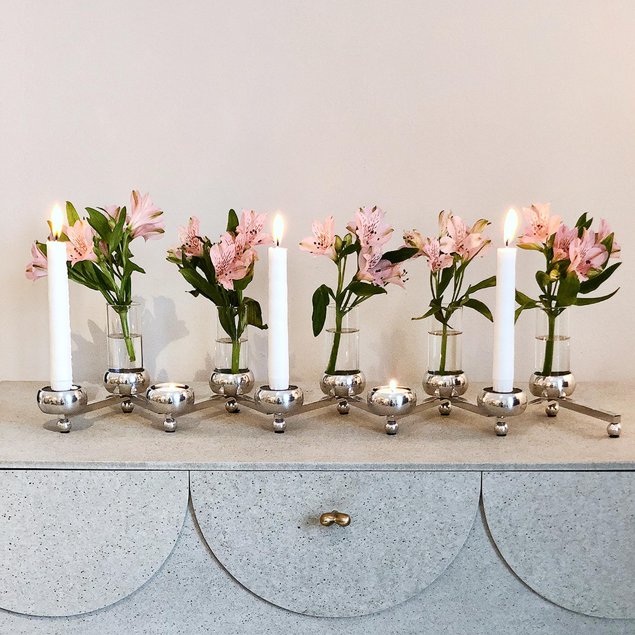 Moderni hopeinen kynttelikkö, jossa on kolme palavaa kynttilää ja pieniä vaaleanpunaisia ​​kukkia sisältävä maljakoita vaaleanharmaalla lipastolla.