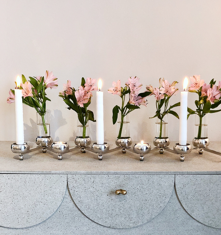 En polerad silverkandelaber med tända vita ljus och rosa alstroemerior i vaser står på en gråspräcklig byrå.