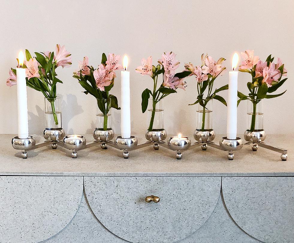 En polerad silverkandelaber med tända vita ljus och rosa alstroemerior i vaser står på en gråspräcklig byrå.