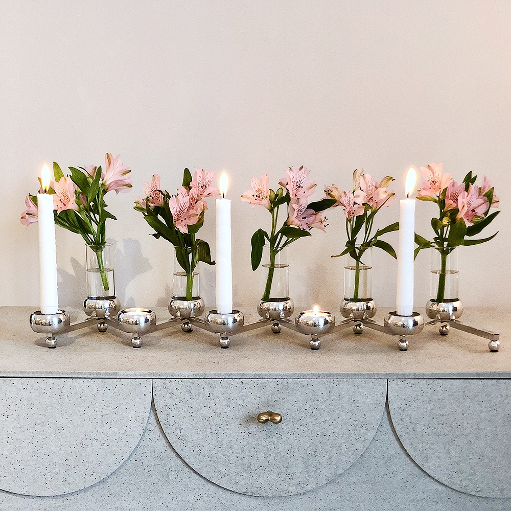 Moderni hopeinen kynttelikkö, jossa on kolme palavaa kynttilää ja pieniä vaaleanpunaisia kukkia sisältävä maljakoita vaaleanharmaalla lipastolla.