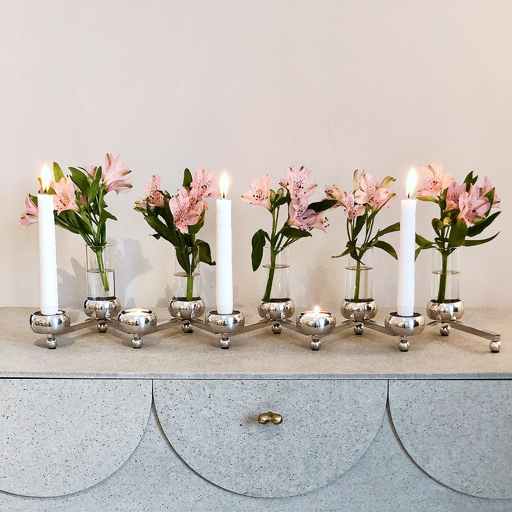 Moderni hopeinen kynttelikkö, jossa on kolme palavaa kynttilää ja pieniä vaaleanpunaisia kukkia sisältävä maljakoita vaaleanharmaalla lipastolla.
